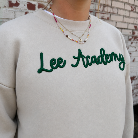 A person wearing a cream sweatshirt with 'Lee Academy' embroidered in dark green script across the chest, paired with layered beaded necklaces, set against a white brick wall background.